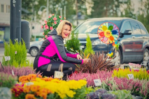 Kuvassa kaunis kukkamyyjä Pieksämäen torilla keltaisten ja punaisten kukkien keskellä hymyilee, taustalla autoja parkkipaikalla.